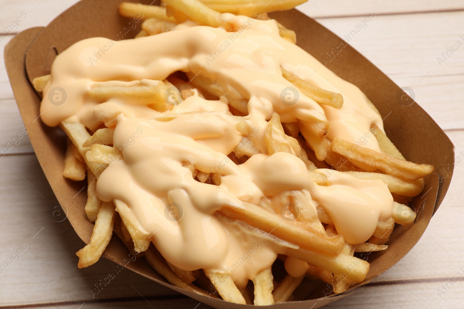 Photo of Tasty potato fries and cheese sauce in paper container on light wooden table, closeup