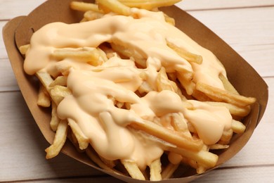 Tasty potato fries and cheese sauce in paper container on light wooden table, closeup