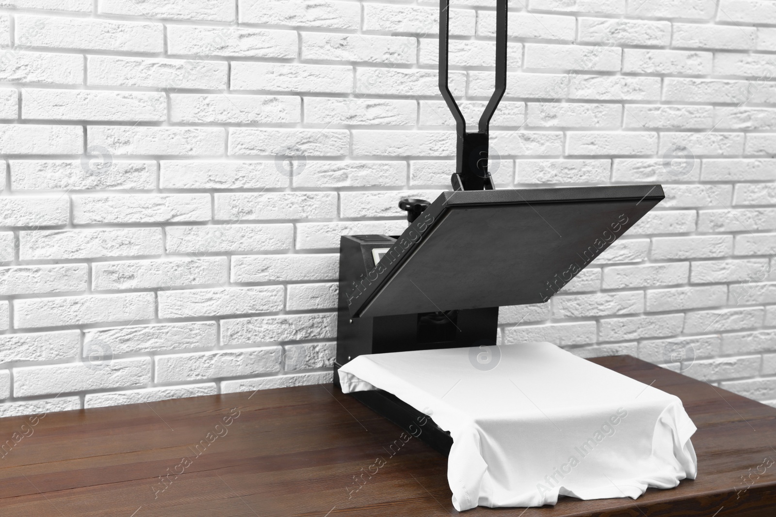 Photo of Heat press machine with t-shirt on wooden table near white brick wall. Space for text