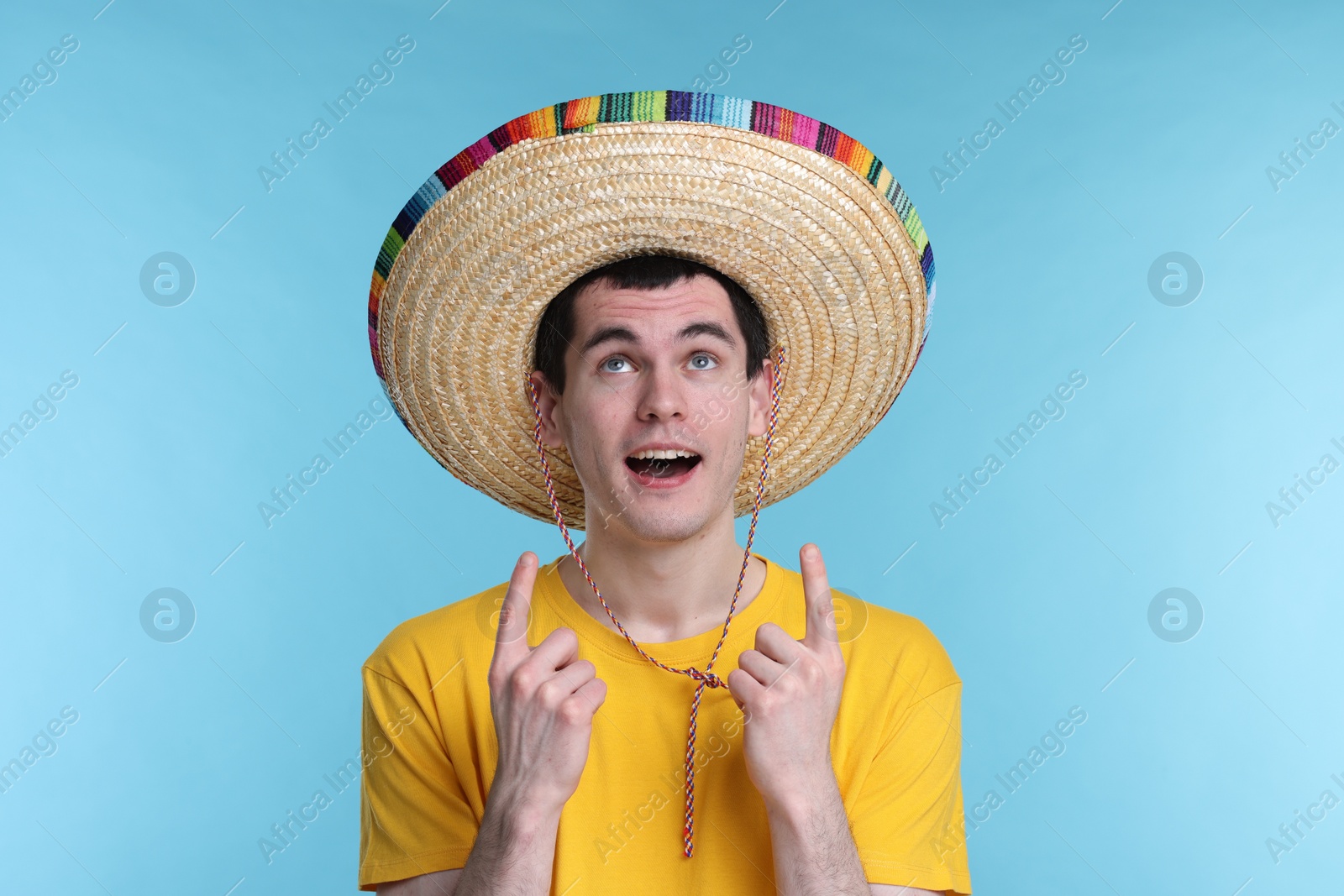 Photo of Young man in Mexican sombrero hat pointing at something on light blue background