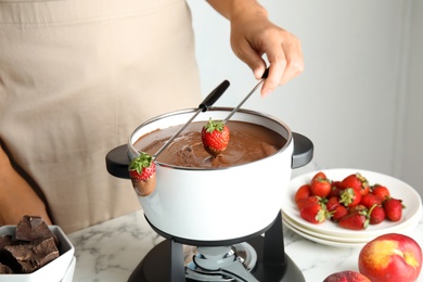 Photo of Woman dipping strawberry into pot with chocolate fondue on table, closeup