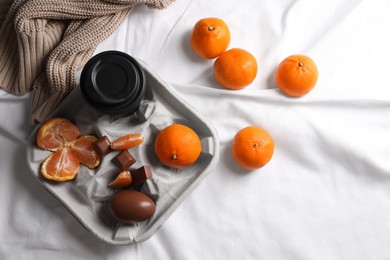 Photo of Delicious ripe tangerines, cup with drink and chocolates on white bedsheet, flat lay