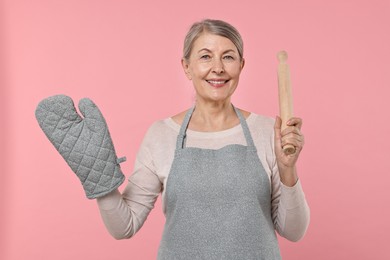 Happy housewife with oven glove and rolling pin on pink background