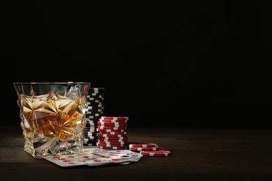Photo of Casino chips, playing cards and glass of whiskey on wooden table, space for text