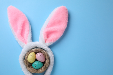 Headband with Easter bunny ears and dyed eggs in nest on light blue background, flat lay. Space for text