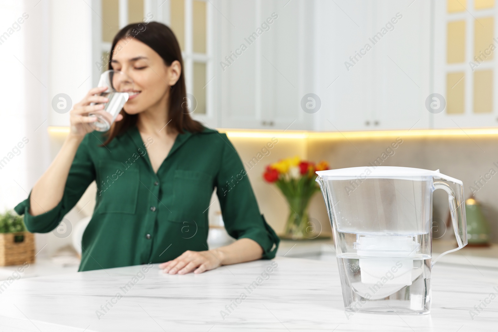 Photo of Woman drinking water in kitchen, focus on filter jug