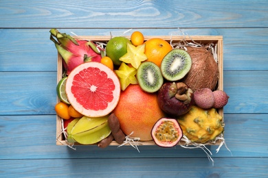 Photo of Different tropical fruits in box on blue wooden background, top view