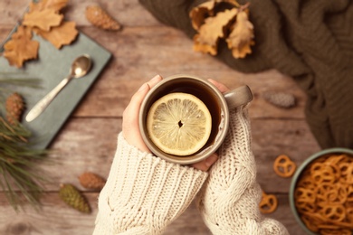 Woman in autumn sweater holding cup of hot cozy drink over wooden table, top view