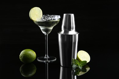 Photo of Metal shaker, delicious cocktail, limes and mint on black mirror surface