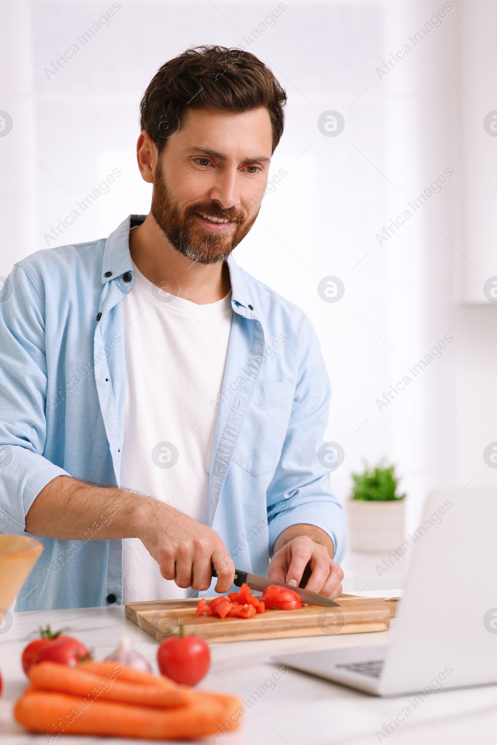 Photo of Man making dinner while watching online cooking course via laptop in kitchen