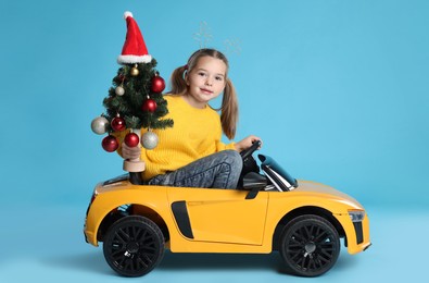 Photo of Cute little girl with Christmas tree driving children's electric toy car on light blue background