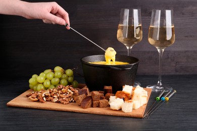 Photo of Woman dipping piece of bread into fondue pot with melted cheese at black wooden table, closeup