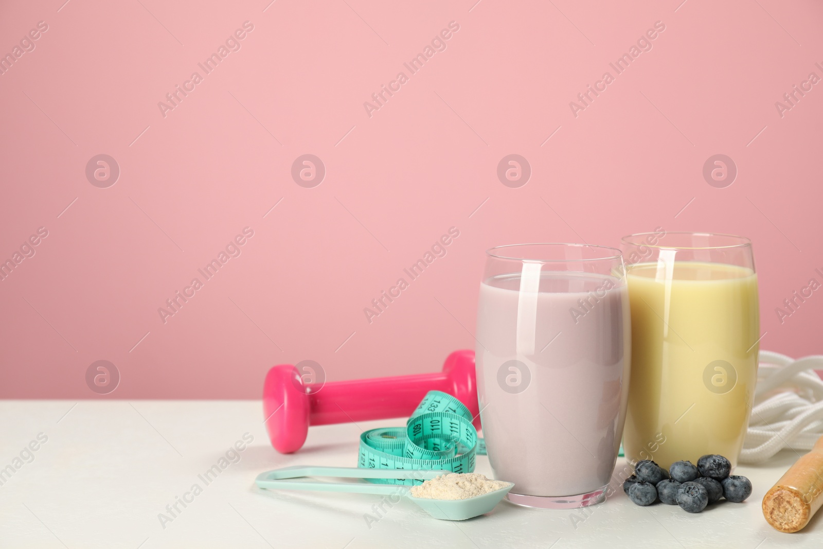 Photo of Tasty shakes with blueberries, sports equipment, measuring tape and powder on white table against pink background, space for text. Weight loss