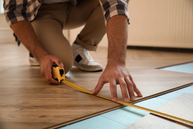 Photo of Professional worker using measuring tape during installation of parquet flooring indoors, closeup