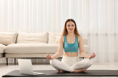 Photo of Happy woman in sportswear meditating near laptop at home. Harmony and zen