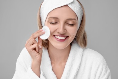 Young woman cleaning her face with cotton pad on light grey background