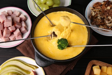 Photo of Dipping different products into fondue pot with melted cheese on black wooden table, above view