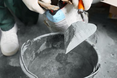 Photo of Worker with cement powder and trowel mixing concrete in bucket indoors, closeup