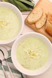 Bowls of tasty leek soup, spoons and bread on white wooden table, flat lay