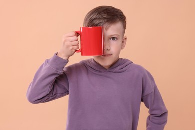 Photo of Cute boy covering eye with red ceramic mug on beige background