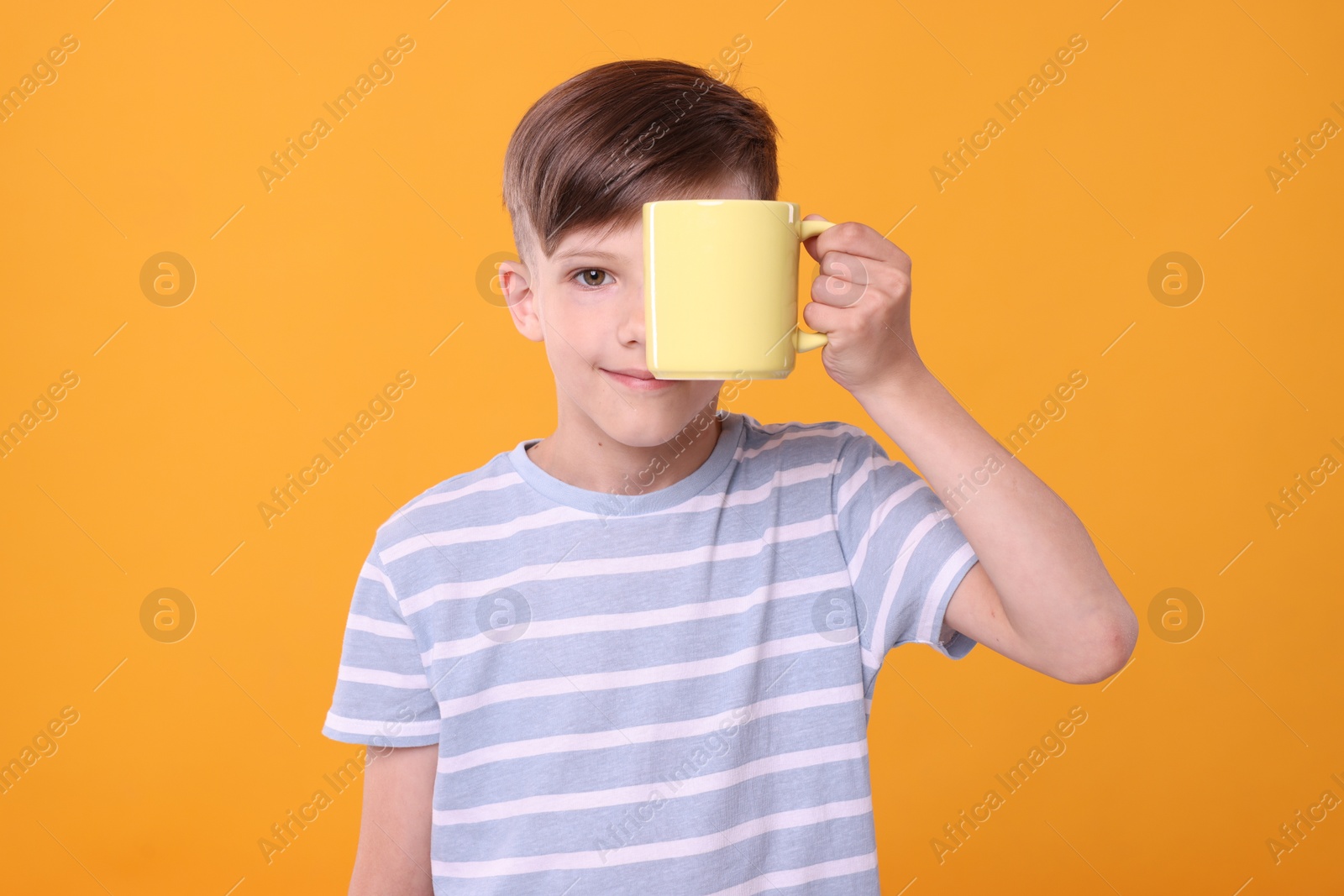 Photo of Cute boy covering eye with yellow ceramic mug on orange background