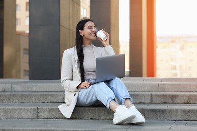 Photo of Happy young woman with cup of coffee using modern laptop on stairs outdoors