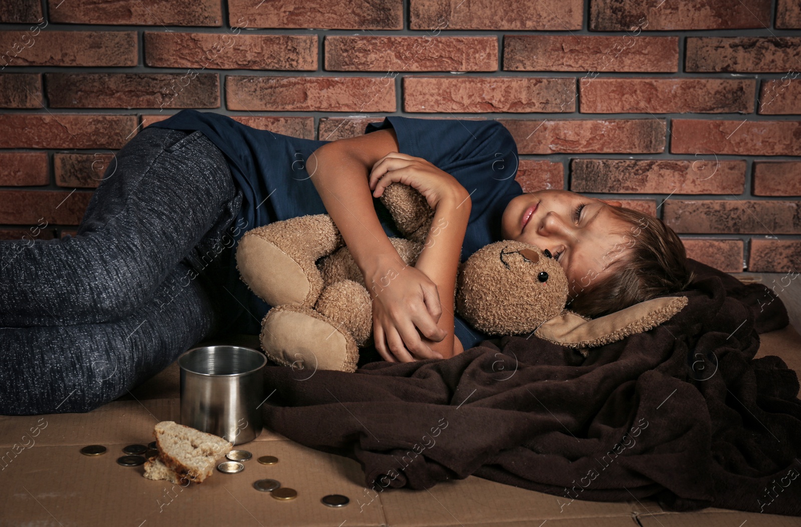 Photo of Poor homeless boy sleeping with his toy on floor near brick wall