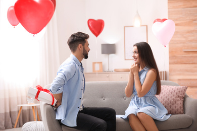 Photo of Young man presenting gift to his girlfriend in living room decorated with heart shaped balloons. Valentine's day celebration