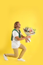 Young handsome man with beautiful flower bouquet bending knee on yellow background