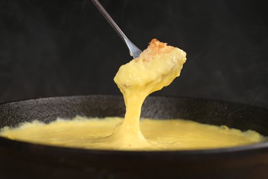 Photo of Dipping piece of bread into fondue pot with tasty melted cheese against dark gray background, closeup