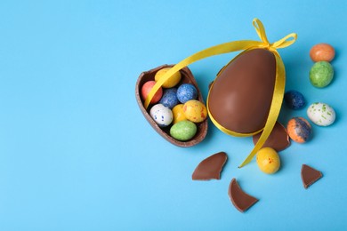 Tasty whole chocolate egg with yellow bow and different candies on light blue background, flat lay. Space for text