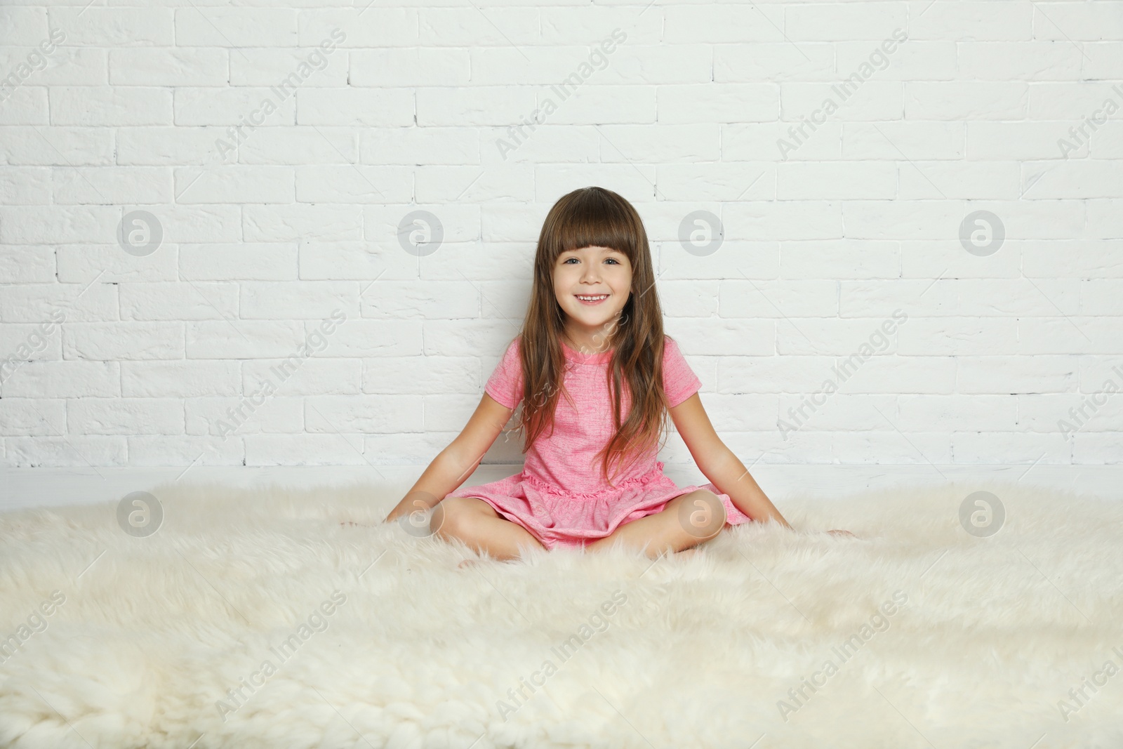 Photo of Cute little girl sitting on fur rug against brick wall