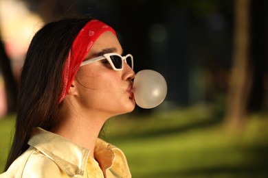 Beautiful young woman in sunglasses blowing bubble gum in park. Space for text