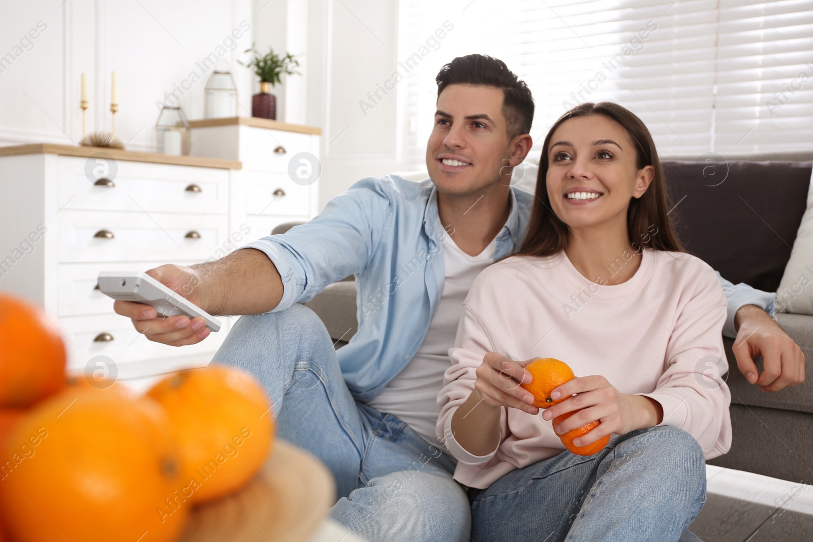 Photo of Happy couple with tangerines and remote control near sofa indoors