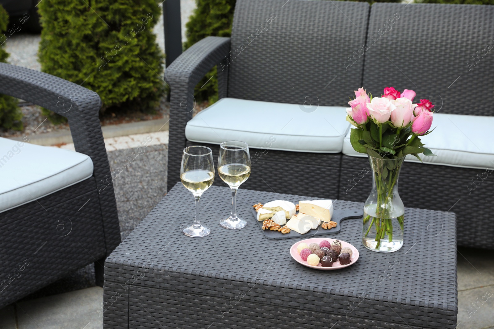 Photo of Vase with roses, glasses of wine and food on rattan table on outdoor terrace