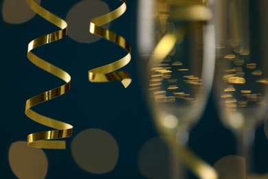 Shiny serpentine streamers and glasses of champagne on dark blue background with blurred lights, closeup