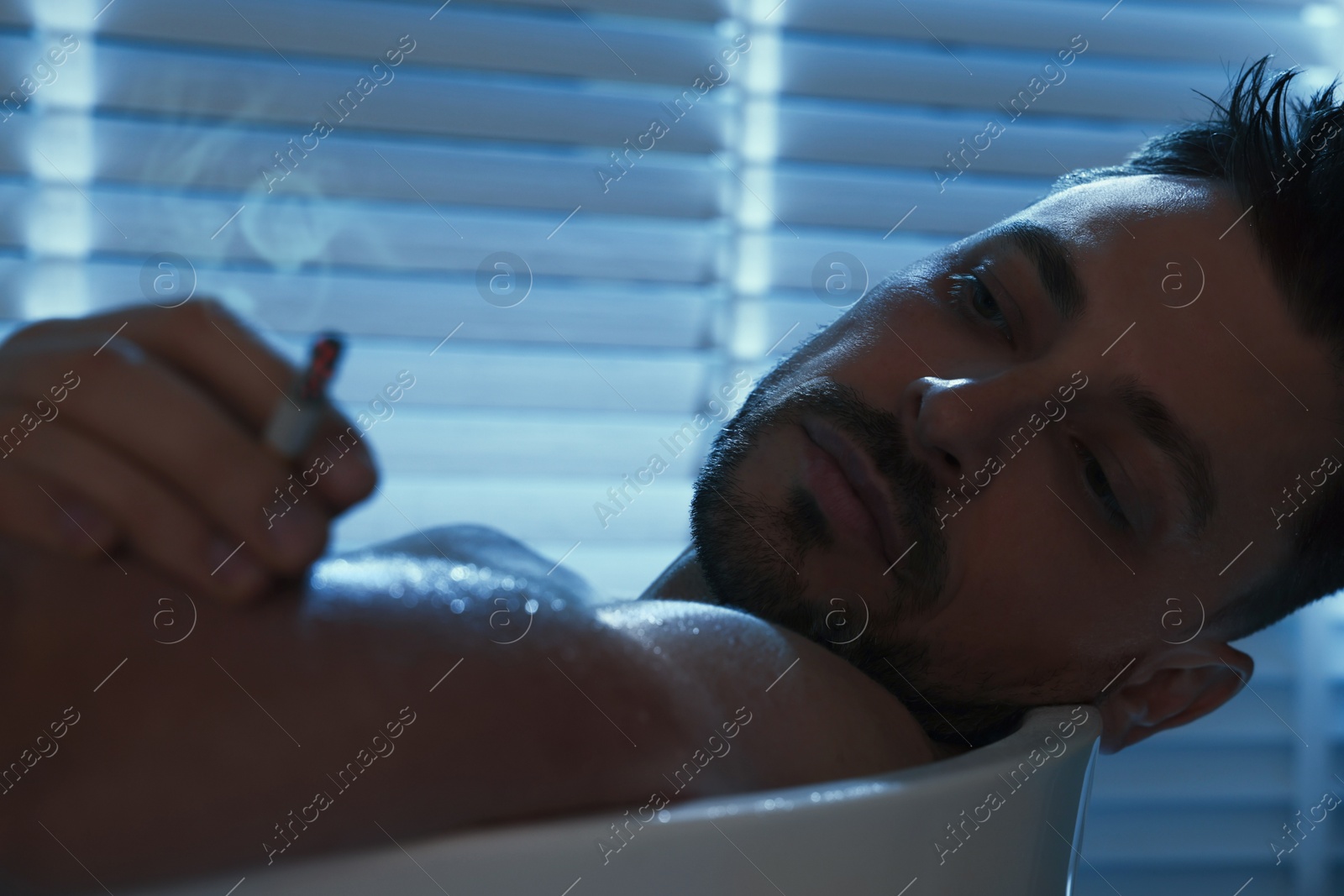 Photo of Upset man smoking in bathtub at night. Loneliness concept