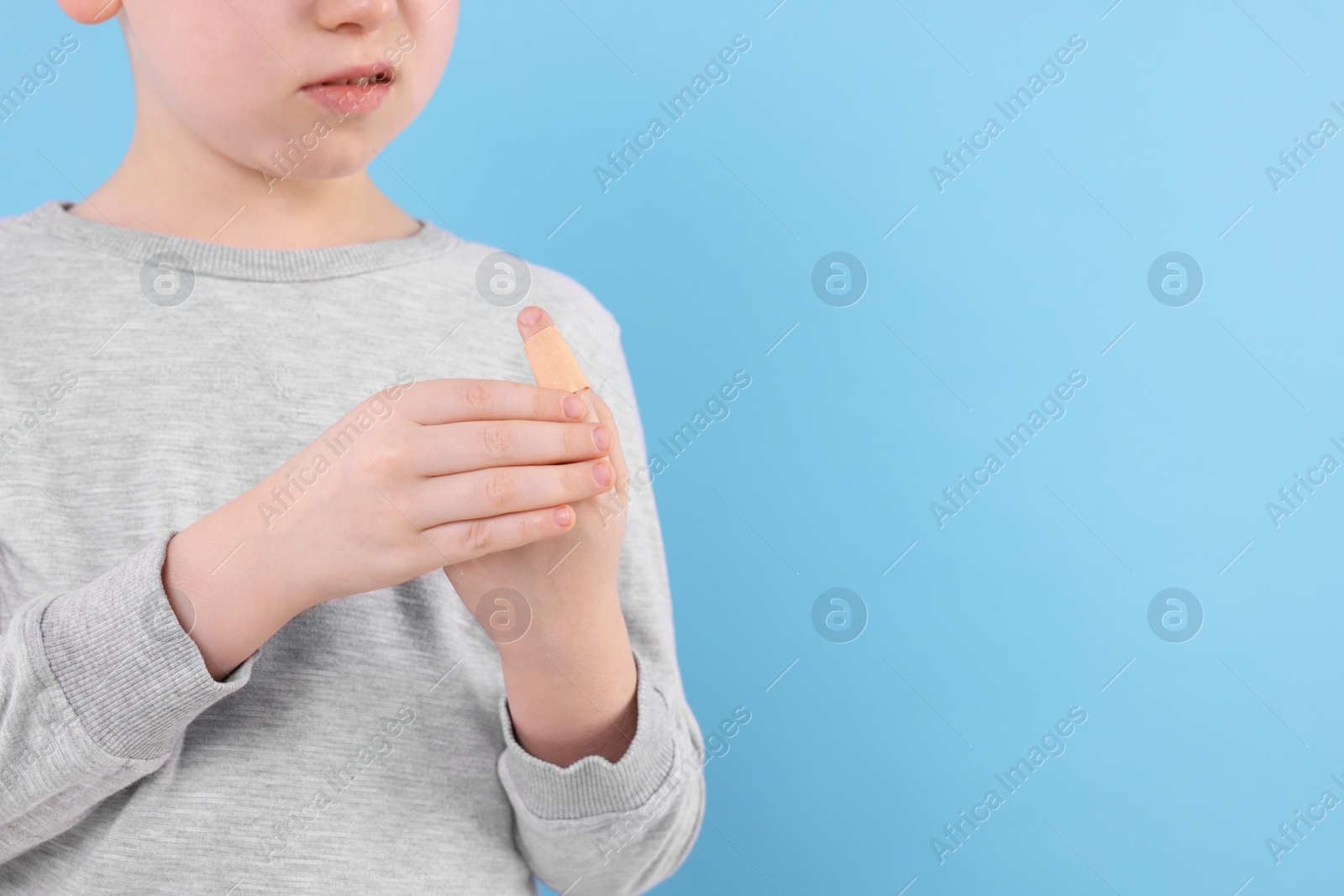 Photo of Little boy putting sticking plaster onto finger against light blue background, closeup. Space for text