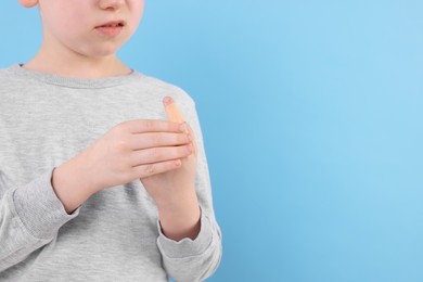 Little boy putting sticking plaster onto finger against light blue background, closeup. Space for text