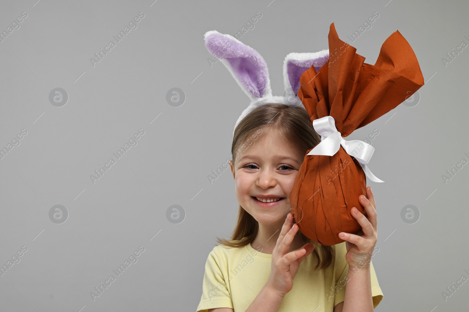 Photo of Easter celebration. Cute girl with bunny ears holding wrapped gift on gray background, space for text