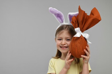 Easter celebration. Cute girl with bunny ears holding wrapped gift on gray background, space for text