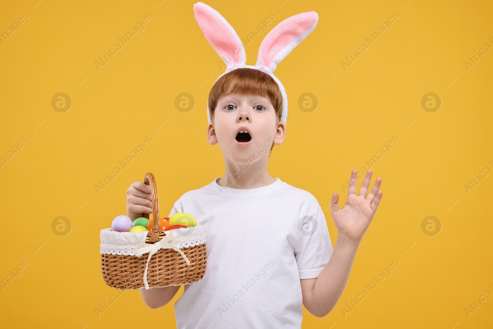 Photo of Easter celebration. Surprised little boy with bunny ears and wicker basket full of painted eggs on orange background