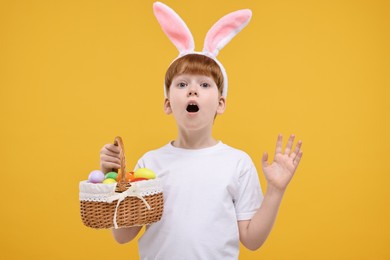 Easter celebration. Surprised little boy with bunny ears and wicker basket full of painted eggs on orange background