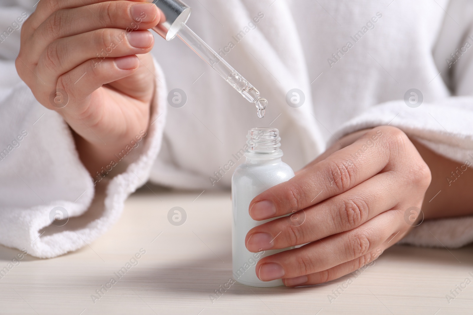 Photo of Woman dripping cosmetic serum from pipette into bottle at white table, closeup