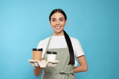 Photo of Young woman in grey apron with hot drinks on light blue background