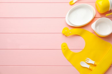 Flat lay composition with silicone baby bib and plastic dishware on pink wooden background. Space for text