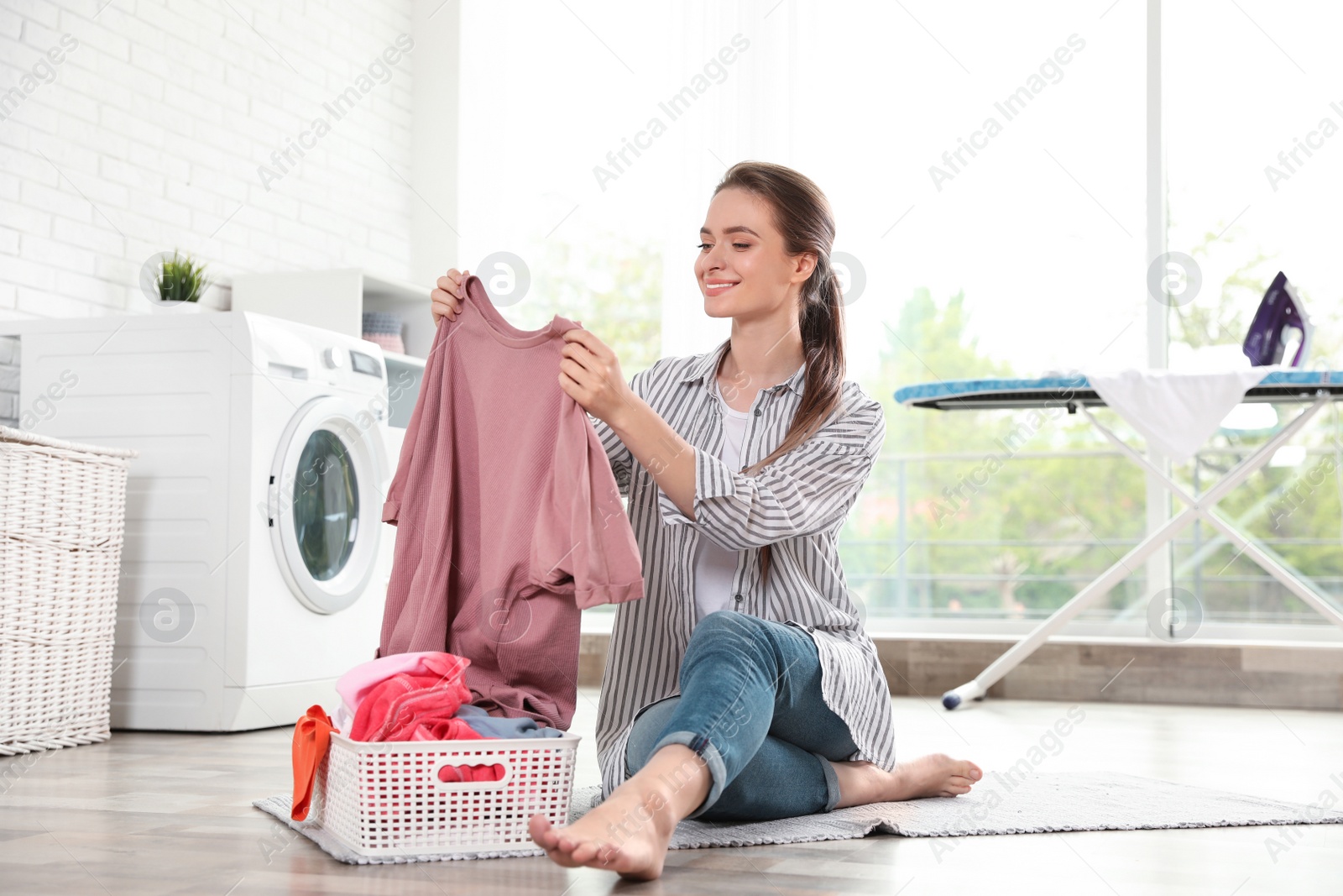 Photo of Young woman with basket of clean laundry on floor in room