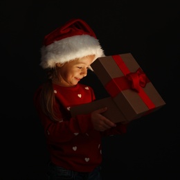 Photo of Cute little girl in Santa hat with Christmas gift box on black background