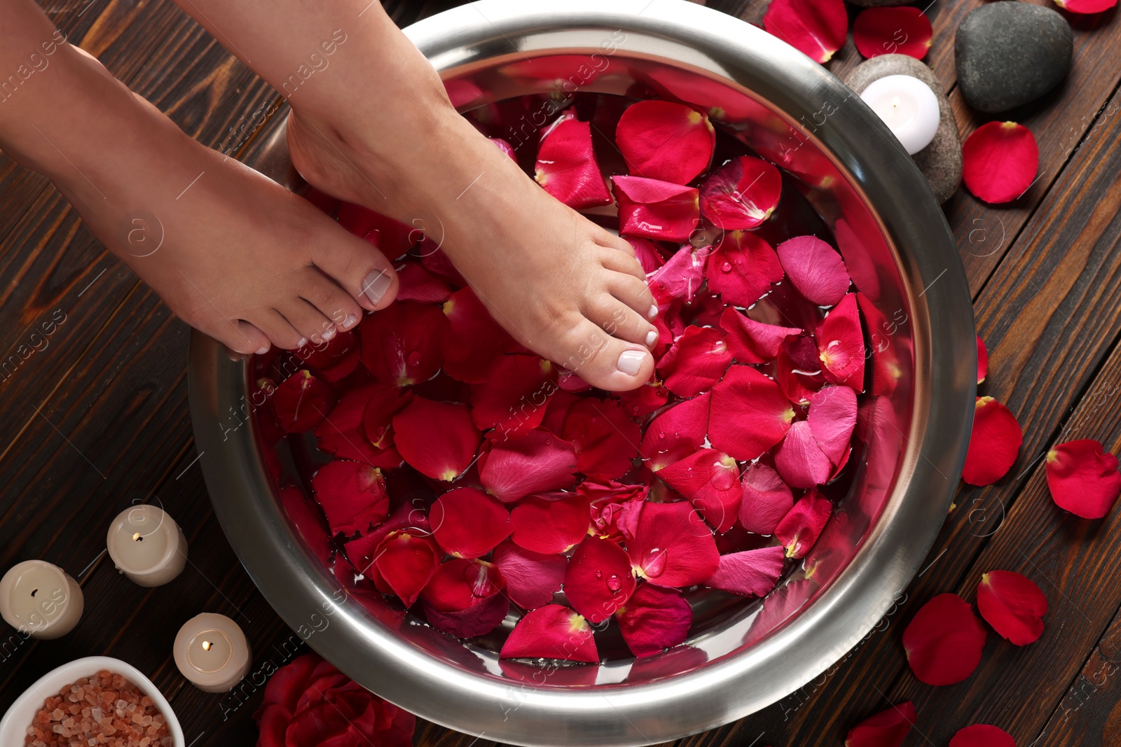 Photo of Woman soaking her feet in bowl with water and red rose petals on wooden floor, top view. Pedicure procedure