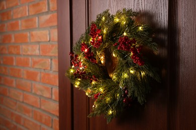 Beautiful Christmas wreath with red berries and fairy lights hanging on wooden door, space for text